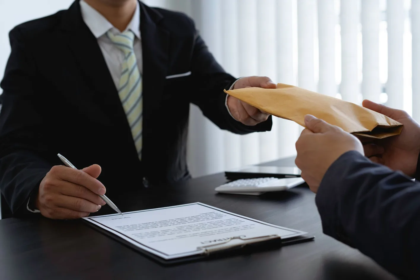 A businessman signing a document as an envelope is passed across the desk