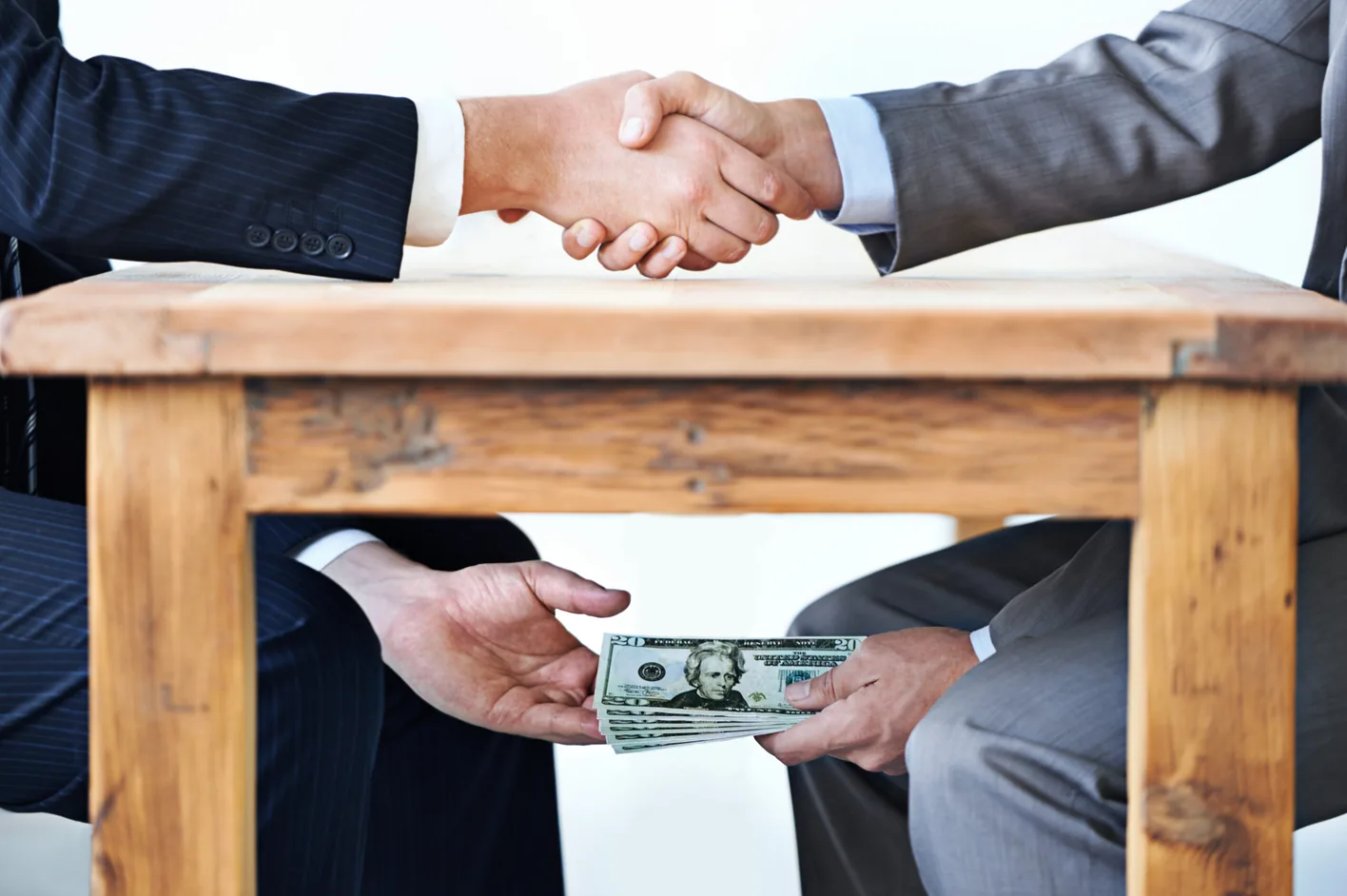 Two businessmen shaking hands over a table while passing cash beneath it
