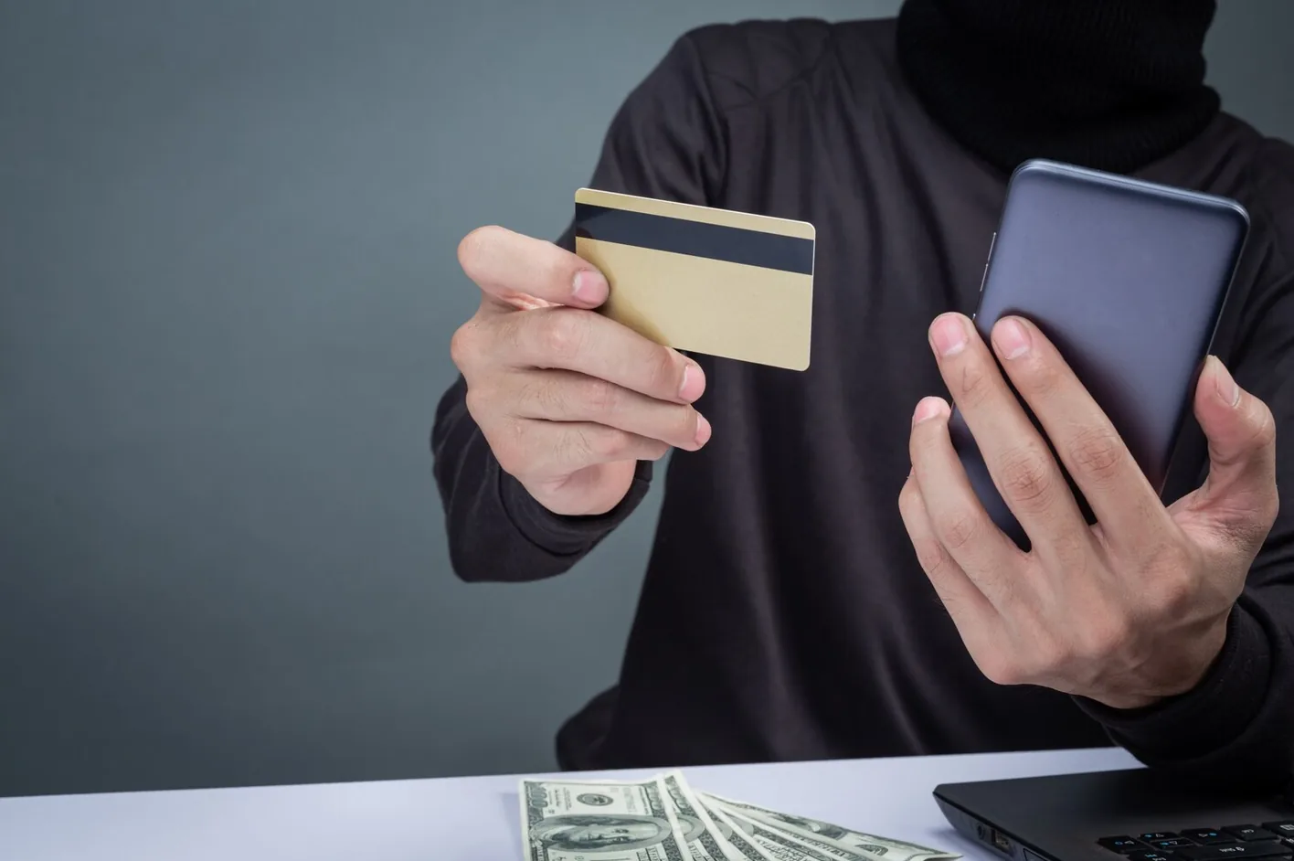 A hooded person holding a credit card and a smartphone at a laptop