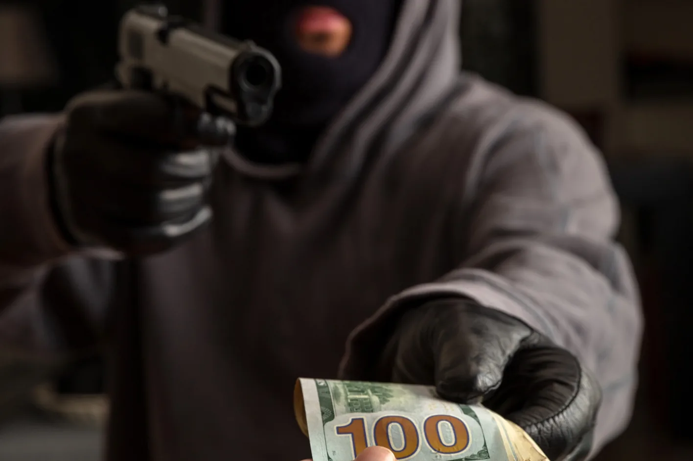 A masked robber pointing a handgun and holding a hundred-dollar bill