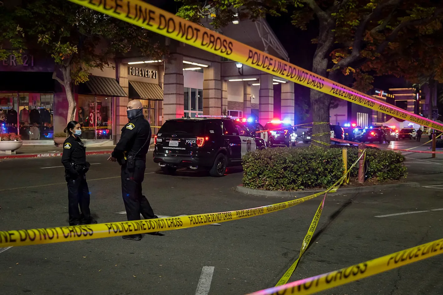 Police officers behind police tape at a night-time scene with patrol cars