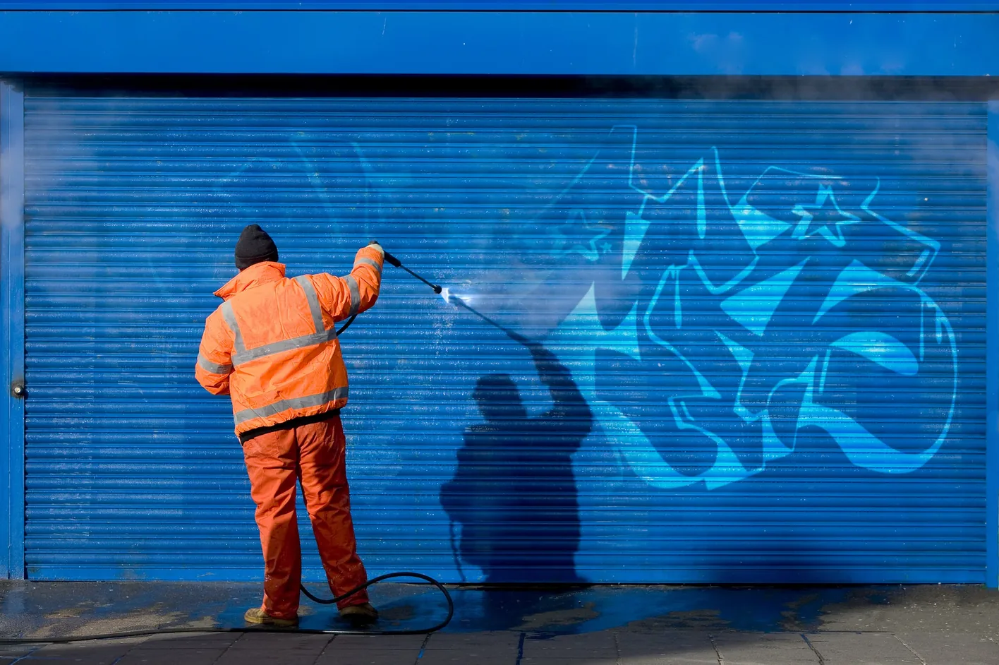 A worker in orange pressure-washing graffiti off a blue shutter