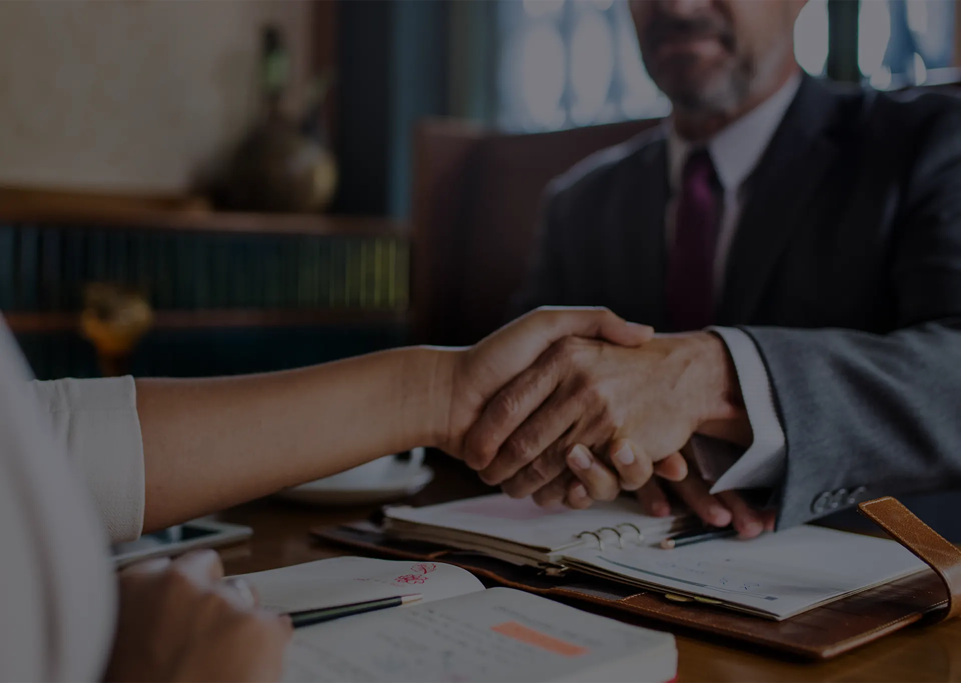 A man in a suit shaking hands with a client across a table, notebooks open between them