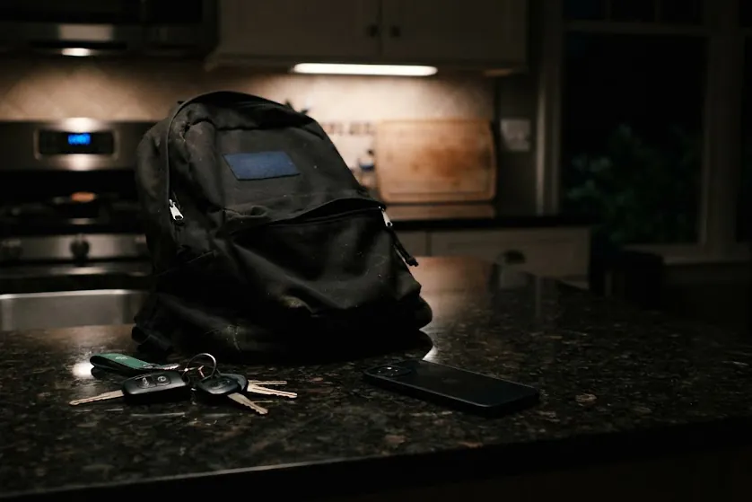Car keys and a school backpack on a kitchen counter at night, a phone face-down beside them